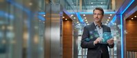 Middle-aged Caucasian male businessman in dark blazer and light blue shirt holding glowing digital tablet in modern glass office corridor with blue lighting and contemporary architecture.