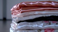 Soft fabric stack of folded bedding and linens in pink, white and floral patterns creating cozy home textile layers with delicate textures and gentle shadows.