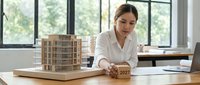 Female architect of mixed ethnicity reviewing architectural model in modern office with large windows, holding 2027 calendar block, representing future planning and design innovation.
