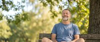 Middle-aged man with light skin tone and short gray hair, wearing casual blue t-shirt, sitting on wooden bench in sunny park surrounded by green trees and natural foliage, relaxed outdoor lifestyle portrait.