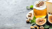 Fresh honey jar with wooden dipper, ripe figs, walnuts and mint leaves arranged on dark stone background. Natural organic ingredients for healthy breakfast.
