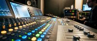 Audio mixing console with illuminated faders and buttons in professional recording studio environment with monitors and equipment in background.