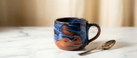 Handcrafted ceramic coffee mug with swirled blue and orange glaze pattern sits on marble surface with golden spoon, perfect for morning beverage rituals.