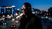 Man wearing sunglasses and black jacket standing against blurred city lights at night, urban lifestyle portrait for advertising and editorial use.