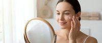 Young woman with East Asian ethnicity applying skincare cream to her face while looking in round wooden mirror, demonstrating beauty routine and cosmetic product use.