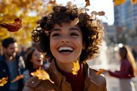 Young African American woman with curly hair laughing joyfully outdoors on autumn day, wearing brown leather jacket, falling yellow leaves and sunlit bokeh background.