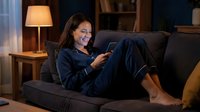 Woman in dark blue pajamas relaxing on couch at night, holding remote control, watching television in cozy living room with warm lamp lighting.