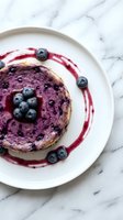 Blueberry cheesecake slice on white plate with fresh berries and sauce drizzle on marble surface for dessert menu and food photography.