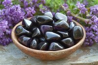 Polished black obsidian stones in rustic ceramic bowl against purple lilac flowers background on weathered wooden surface, spiritual healing crystals display.