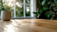 Sunlit wooden table surface with soft natural light streaming through window, creating gentle shadows. Small potted plant adds organic element to minimal scene.