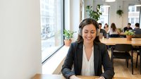Professional businesswoman wearing headset smiling while working in modern office environment with colleagues in background for customer service concepts.