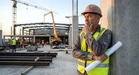 Middle-aged Caucasian construction foreman in hard hat and safety vest holding blueprints at active building site with crane and workers in background.