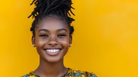 Young African woman with dreadlocks hairstyle and traditional print clothing smiling brightly against vibrant yellow background, showing white teeth and natural joy.