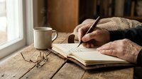 Elderly hands writing in notebook at wooden table with coffee cup and glasses, cozy morning journaling scene for retirement lifestyle content.