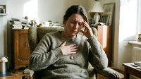 Stressed young woman experiencing anxiety attack touching chest and head while sitting on couch at home during mental health crisis.