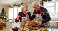 Happy senior Caucasian couple preparing Christmas turkey dinner together in festive kitchen wearing matching aprons and holiday sweaters.