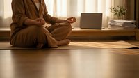 Woman in brown robe practicing meditation in lotus position on wooden floor near laptop in warm sunlit room creating peaceful mindfulness workspace atmosphere.