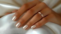 Elegant female hand with pearl white manicure and gold wedding band resting on soft ivory satin fabric, showcasing modern bridal style.