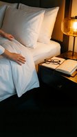 Cozy bedroom interior with hands adjusting white bedding, eyeglasses and book on nightstand, warm ambient lighting for hospitality and comfort concepts.