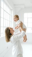 Happy Caucasian mother in white robe lifting laughing blonde toddler in bright modern home interior with large windows for family lifestyle content.