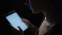 Young woman using tablet computer in dark room with glowing screen light illuminating her face for late night browsing and digital technology concepts.