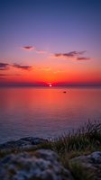 Stunning sunset over calm ocean waters with vibrant orange and purple sky, small boat on horizon, rocky shore and coastal vegetation in foreground. Perfect for travel and nature projects.