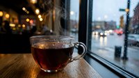 Steaming hot tea cup on wooden table by rainy window with blurred city lights and wet street reflections creating cozy cafe atmosphere for relaxation.