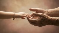 Young woman's hand reaching toward elderly person's weathered hands in warm sunlight representing intergenerational connection and care.