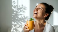 Young woman of South Asian descent enjoying fresh orange juice, smiling with natural sunlight casting leaf shadows on white wall background.