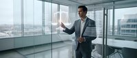 Professional businessman in modern office presenting data on transparent screen with city skyline view through floor-to-ceiling windows for corporate presentations.