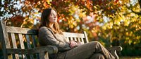 Young woman relaxing on park bench during golden autumn day, wearing cozy sweater and enjoying peaceful moment outdoors in warm sunlight.