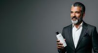 Mature man with salt-and-pepper beard and dark hair, appearing to be in his 50s of Mediterranean or Middle Eastern descent, wearing a charcoal business suit with white dress shirt, holding a white cosmetic bottle against neutral gray background for skincare, grooming or men's product advertising.