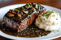 Grilled beef steak topped with sauteed mushrooms and rich brown sauce, served with creamy mashed potatoes garnished with fresh herbs on white plate.