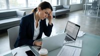 Stressed businesswoman with headache working on laptop in modern office with financial documents and reports on desk.
