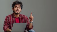 Young South Asian male professional in casual red plaid shirt holding laptop computer while pointing finger up showing idea or solution against gray background.