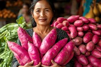 Asian female vendor smiling while holding fresh purple sweet potatoes at local produce market, surrounded by colorful vegetables and tropical fruits.