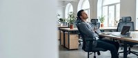 Relaxed businessman stretching in ergonomic office chair during work break in modern workspace with laptop and plants for stress relief and wellness.