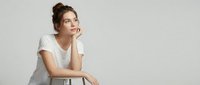Young thoughtful woman in white t-shirt sitting on chair with hand on chin, looking away pensively against neutral background for lifestyle content.