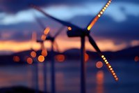 Wind turbines silhouetted against dramatic sunset sky with bokeh lights along blades, reflecting on water surface creating atmospheric renewable energy scene.