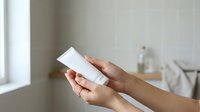 Person with medium skin tone holding white cosmetic product container in bathroom setting for skincare, beauty, and wellness marketing.