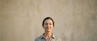 Middle-aged woman with dark hair and fair complexion wearing casual gray shirt against neutral beige textured wall background, suitable for corporate portraits, business profiles, and professional headshots.