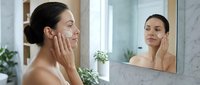 Young woman with dark hair and olive complexion applying skincare product to face while looking in bathroom mirror, demonstrating daily beauty routine and facial treatment application.