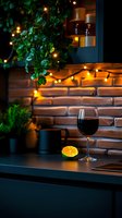 Glass of red wine with orange slice on dark counter against brick wall background with warm string lights and green plants creating cozy atmosphere.