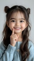 Young Asian girl with space buns hairstyle wearing light blue sweater shows playful expression with finger on smiling lips, captured against neutral background.