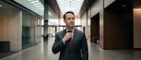 Professional businessman in dark suit adjusting tie while walking through modern office corridor with glass walls and contemporary architecture.