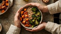 Overhead view of hands holding a bowl with roasted sweet potato, broccoli, and quinoa grain bowl for healthy eating and meal prep content.