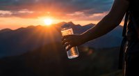 Hiker holding water bottle during golden hour mountain sunset with dramatic sky and silhouetted peaks creating inspirational outdoor adventure scene.