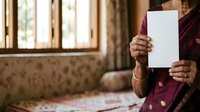 Senior woman with South Asian ethnicity wearing traditional maroon attire holding blank white paper indoors with window and curtains in background for mockup and advertising purposes.
