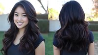 Young Asian woman with long wavy dark hair and bright smile wearing black top shown from front and back view, demonstrating modern hairstyle against blurred outdoor background.