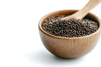 Whole black peppercorns in wooden bowl with spoon on white background, aromatic spice and seasoning ingredient for cooking and food preparation.
